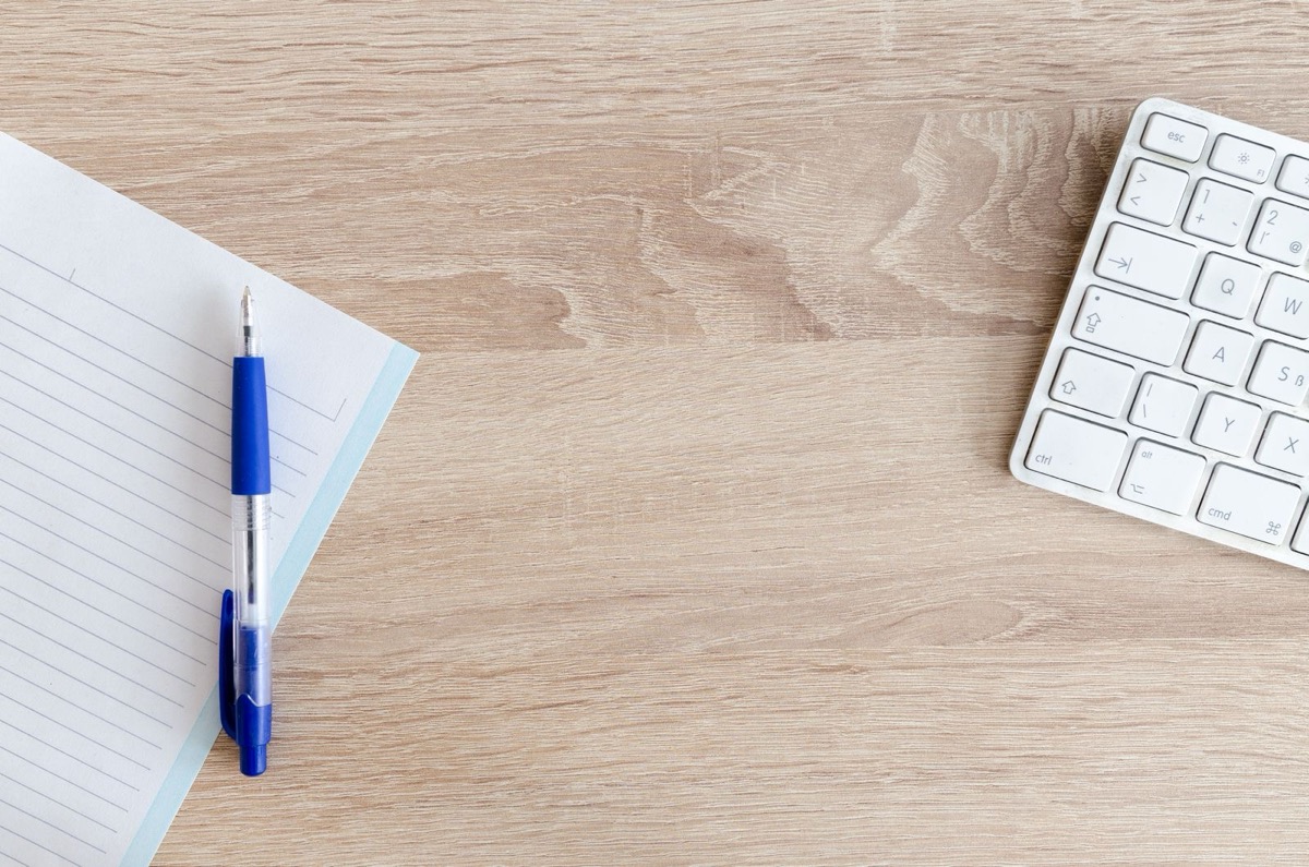 A lined notepad, a pen and a keyboard on a wooden desk