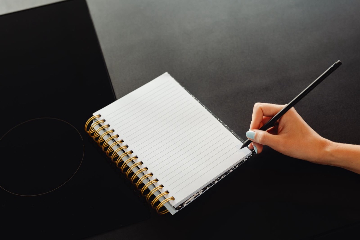 A hand writing in a spiral notebook on a dark surface