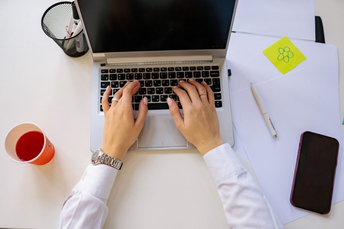 Hands typing on a laptop beside papers, a phone and a sticky note