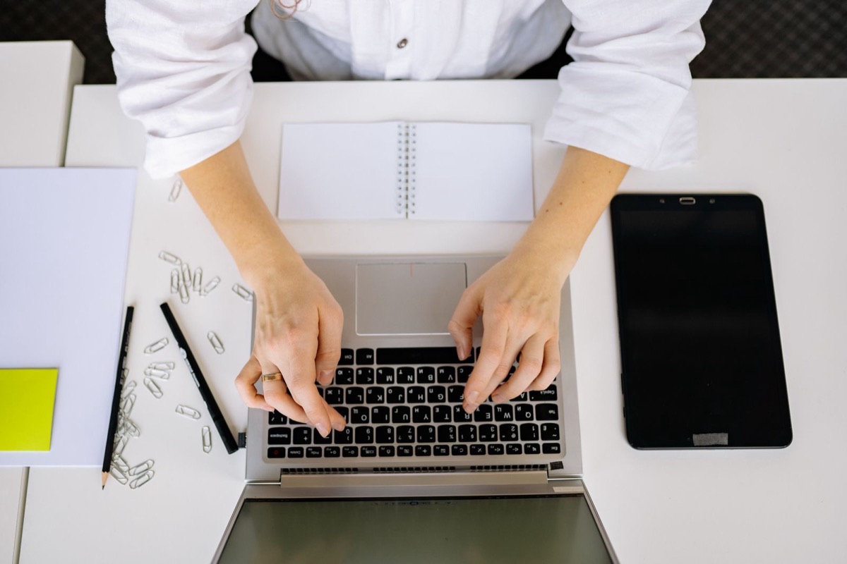 Hands typing on a laptop beside a notepad, a tablet and paper clips