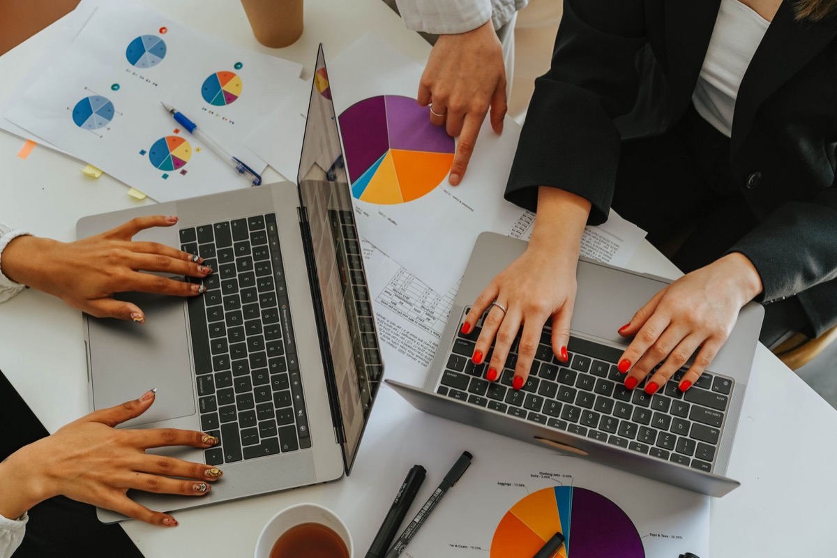 Hands at two laptops among printed pie charts on a white table