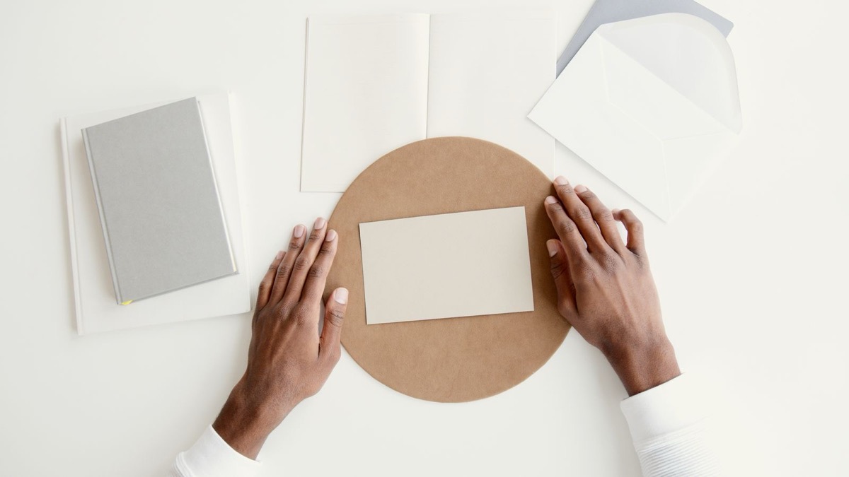 Hands laying a blank card on a desk beside envelopes and a notebook